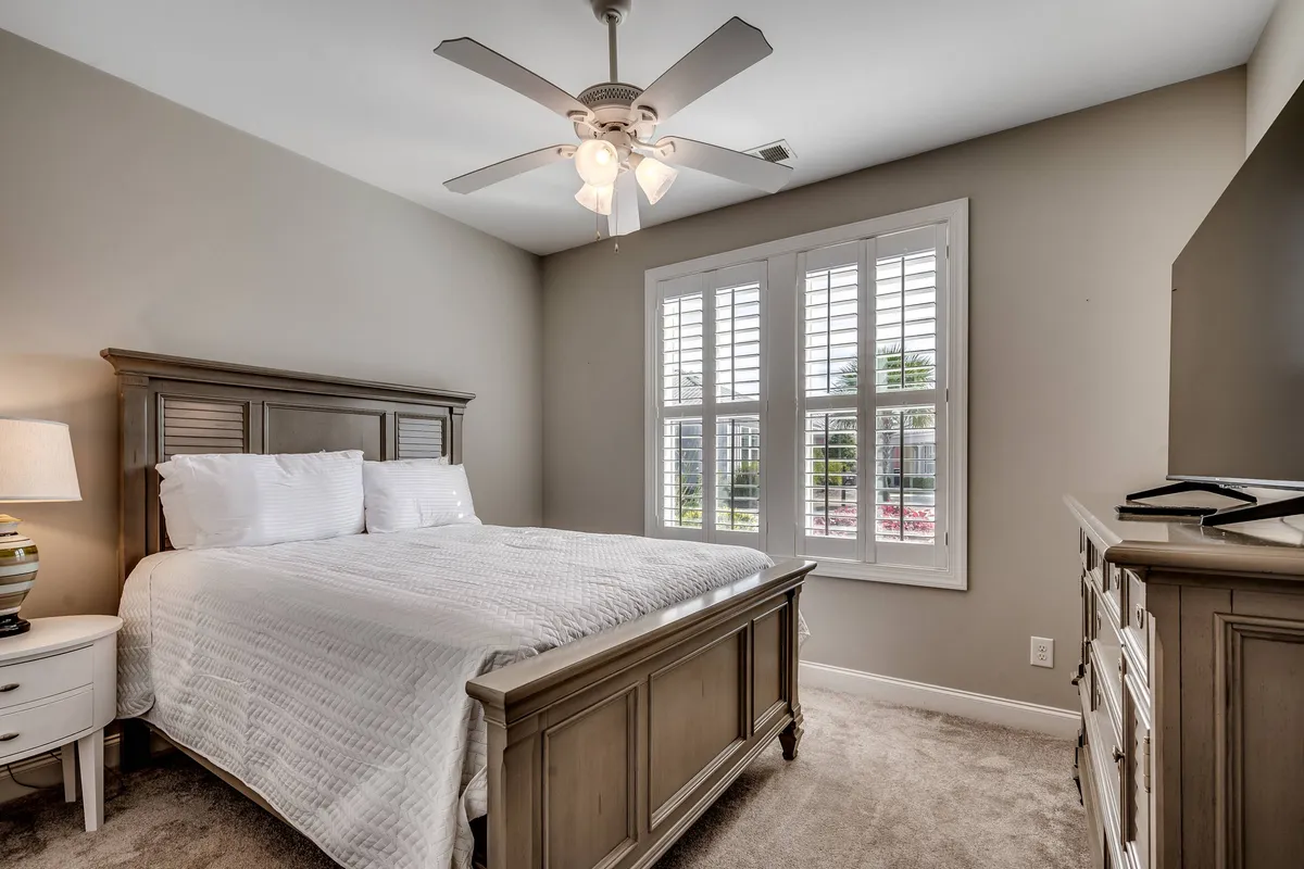 A cozy bedroom featuring a comfortable bed with a nightstand, a window that lets in natural light, a dresser, and a ceiling fan. The setup is tailored for rest and relaxation, with the window offering the possibility of enjoying views outside or a beautiful sunset.