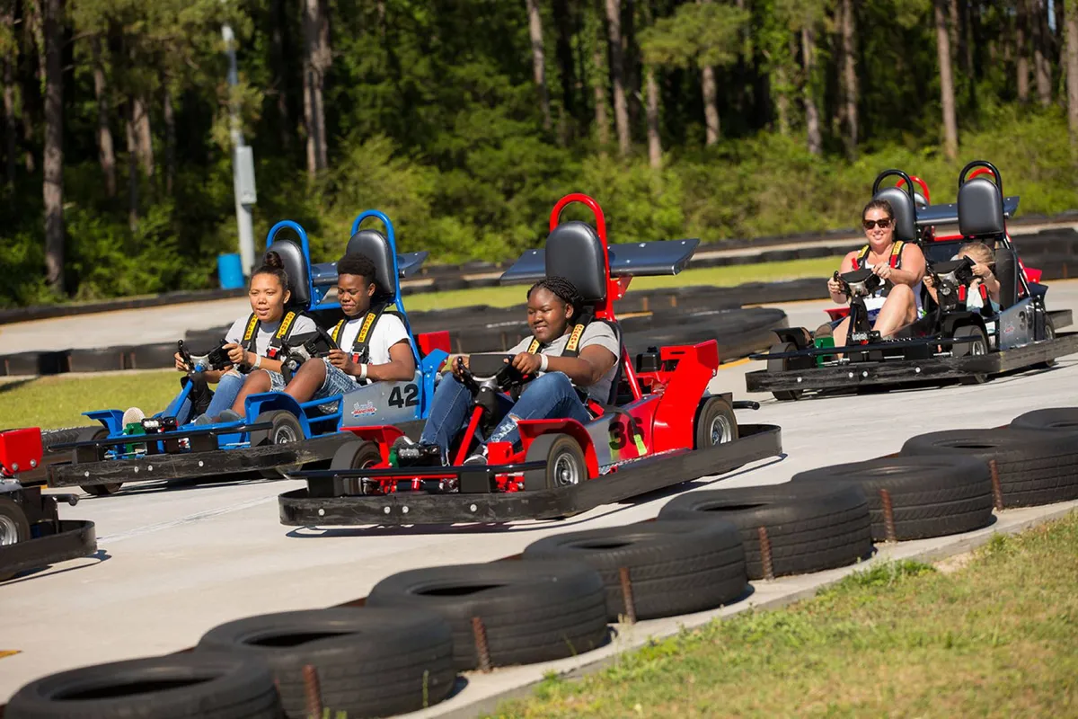 An outdoor scene featuring a go-kart race track bustling with participants steering their karts at speed. The area is surrounded by lush greenery, and the energy of families and friends enjoying competitive racing fills the atmosphere.