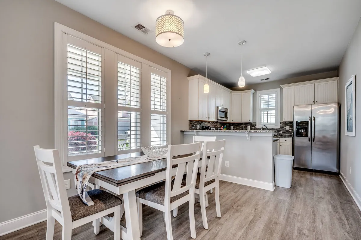 A welcoming shot focused on a dining table and kitchen island, framed by windows with shutters that let in natural light. The scene suggests a space for intimate family meals or festive gatherings, with easy access to the kitchen for preparing and sharing food.