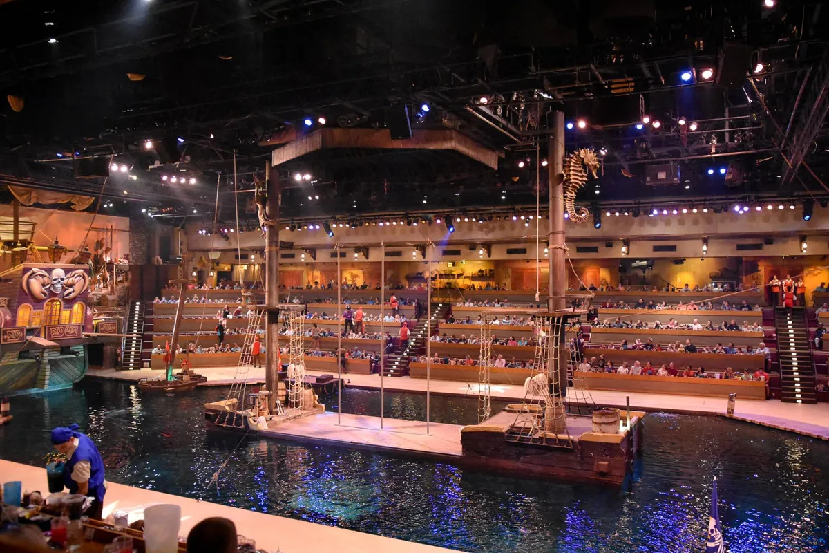 An interior view of a live performance venue, showing a stage with water elements, audience seating, and performers with props, creating an atmosphere of excitement and entertainment. The decorative ship elements suggest a themed show, enhancing the sense of spectacle and memorable group experiences.