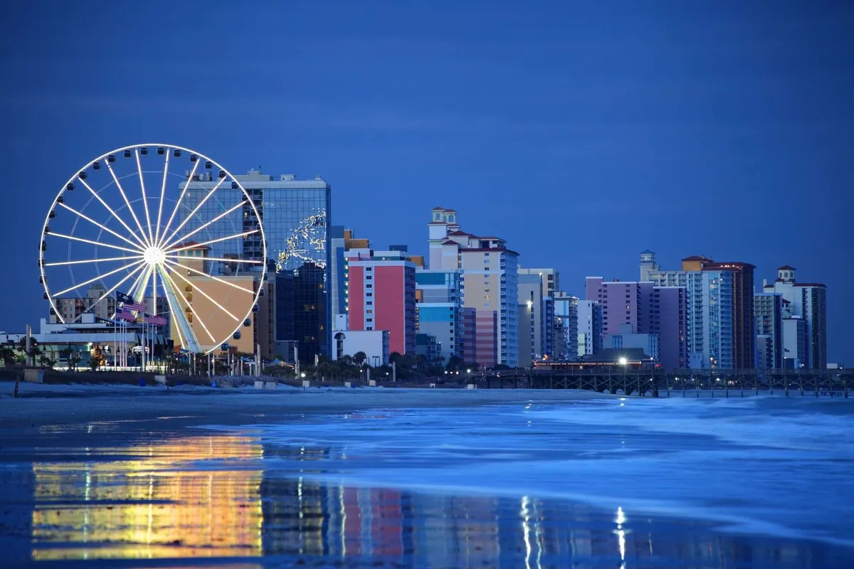 A vibrant exterior shot showcasing a large ferris wheel set against colorful buildings and beautiful ocean reflections. The scene is lively, evoking a festive mood with people enjoying amusement rides, beach walks, and dining by the seaside.