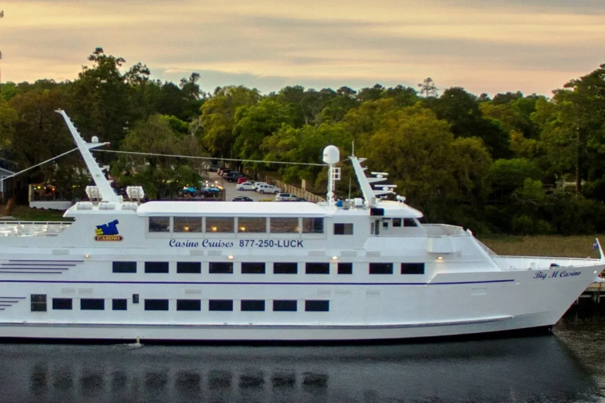 The image displays the exterior of a cruise ship surrounded by lush trees and calm water. The setting sun casts a warm glow, and guests are visible dining on the deck, enjoying the festive ambiance. The scene evokes a feeling of a relaxed, scenic getaway with gentle breezes and opportunities for quiet moments or conversation.