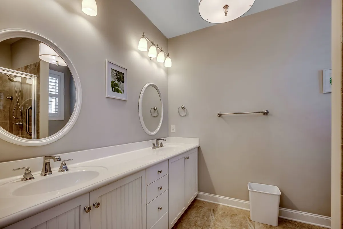 A spacious bathroom with a double sink vanity, large round mirrors, elegant lighting, and framed artwork, evoking a calm and sophisticated spa-like feel. The design encourages relaxation and self-care routines.