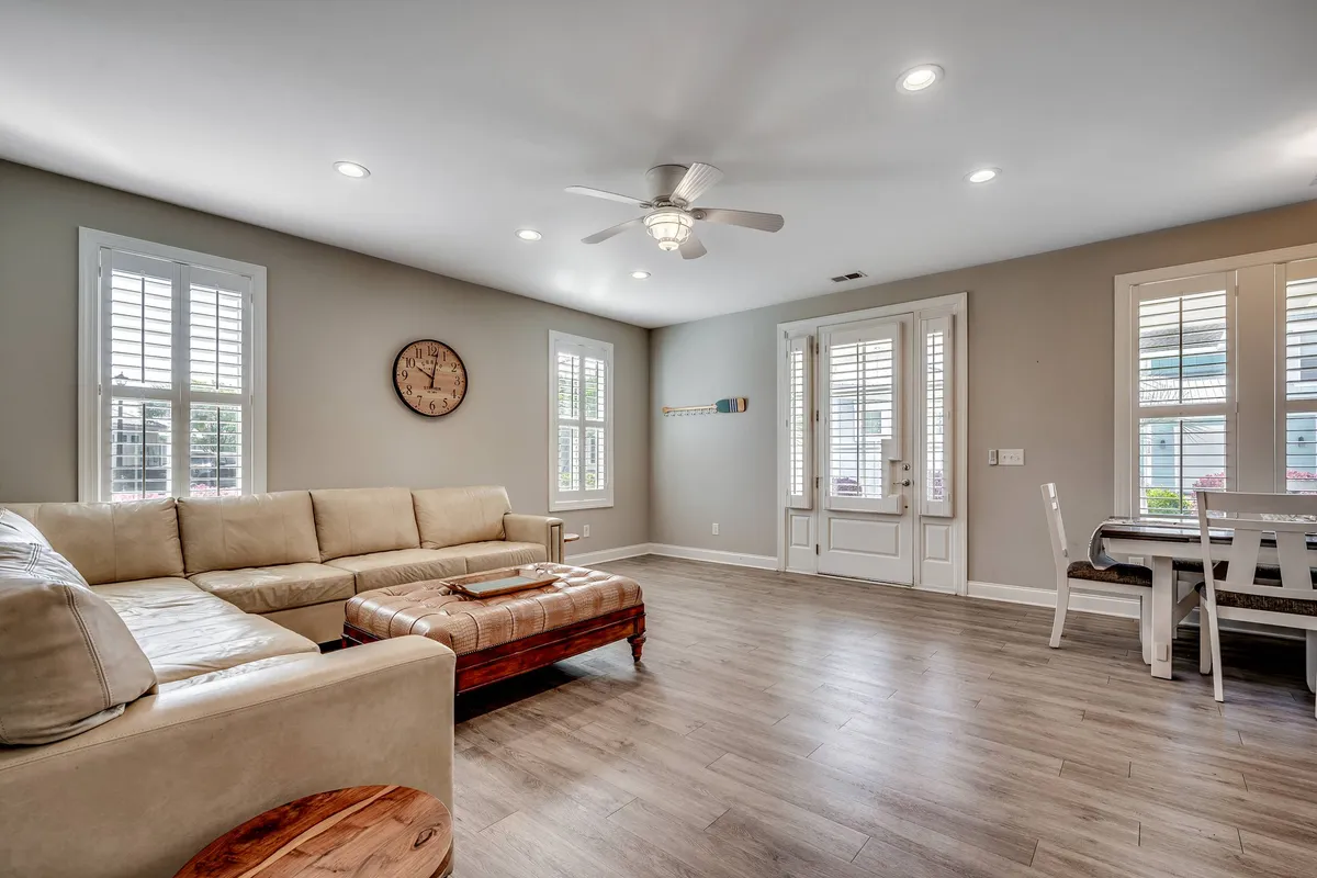 A cozy living room view with a large sectional sofa and wooden coffee table, adjacent to a dining table and chairs. Decor elements like a wall clock add character, and large windows suggest a bright, inviting atmosphere for gatherings or lounging after a day at the beach.