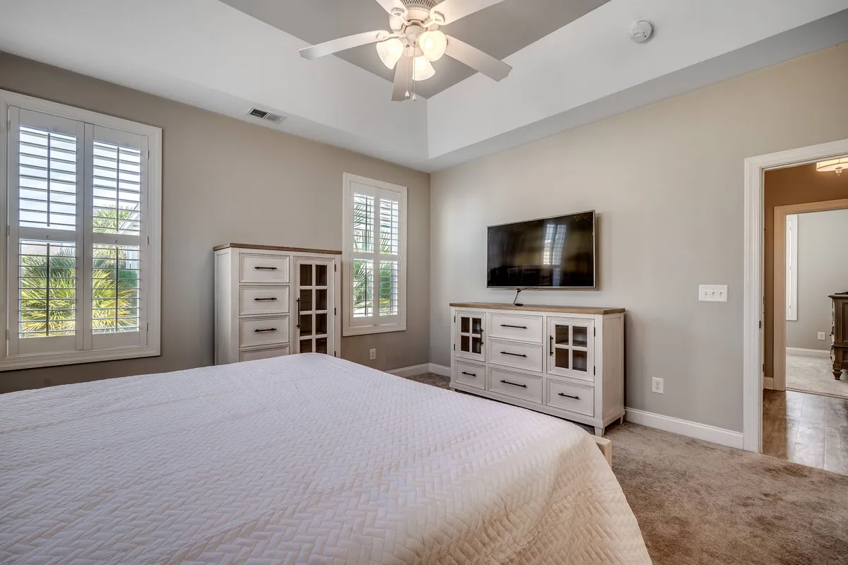 A bright bedroom with a bed dressed in crisp white linens, a dresser with glass doors, a TV mounted on the wall, and windows with shutters that allow sunlight to stream in. The room feels cozy and modern, perfect for relaxation or a cozy movie night.