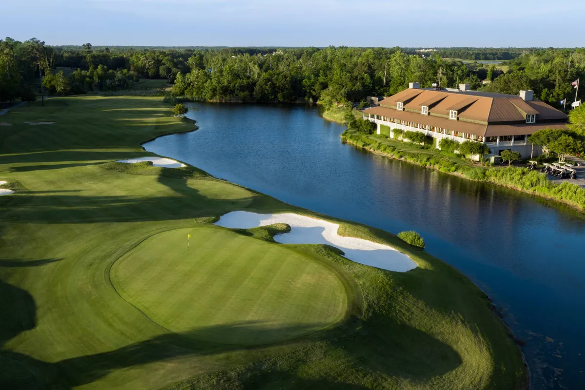 A lush, green golf course stretches out beside a shimmering water feature, with a clubhouse visible in the background and sand traps accenting the fairways. The scene is serene, suggesting both recreation and relaxation in a scenic setting.
